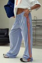 Load image into Gallery viewer, Model wearing powder blue velour-textured wide-leg sweatpants with orange triple side stripes, elastic drawstring waistband, and relaxed oversized fit.