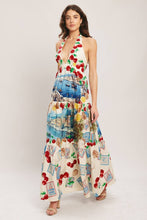 Load image into Gallery viewer, Model wearing a cream halter maxi dress featuring colorful Mediterranean coastal scenes, cherries, lemons, and postcard-inspired prints with a flowing tiered silhouette.
