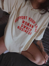 Load image into Gallery viewer, Cream 100% cotton graphic T-shirt with bold red lettering reading "Support Your Local Woman-Owned Business," shown on a model wearing the relaxed-fit tee.