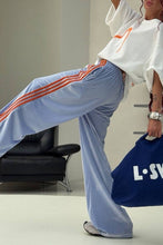 Load image into Gallery viewer, Side view of powder blue velour sweatpants highlighting three orange racing stripes running down the wide legs.