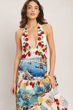 Load image into Gallery viewer, Model wearing a cream halter maxi dress featuring colorful Mediterranean coastal scenes, cherries, lemons, and postcard-inspired prints with a flowing tiered silhouette.