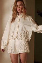 Load image into Gallery viewer, Model wearing an écru cropped shirt jacket with oversized floral appliqué embroidery and matching neutral tailored shorts in a soft resort-inspired setting.