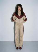 Load image into Gallery viewer, Front view of a female model wearing the Retro Varsity beige and burgundy two-piece track set, featuring a chevron colorblock zip-up jacket and matching high-rise straight-leg pants.