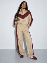 Load image into Gallery viewer, Front view of a female model wearing the Retro Varsity beige and burgundy two-piece track set, featuring a chevron colorblock zip-up jacket and matching high-rise straight-leg pants.