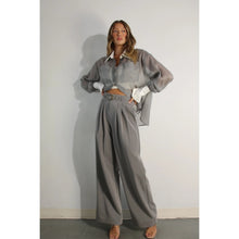 Load image into Gallery viewer, A model showcases a pair of high-waisted, grey pleated trousers, complete with a matching belt, exuding a timeless and sophisticated aesthetic. The trousers feature a tailored fit with front pleats and a straight-leg silhouette, offering both comfort and elegance. Paired with a sheer organza blouse, the ensemble presents a harmonious blend of textures and styles, perfect for both professional and formal occasions.
