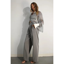 Load image into Gallery viewer, A model showcases a pair of high-waisted, grey pleated trousers, complete with a matching belt, exuding a timeless and sophisticated aesthetic. The trousers feature a tailored fit with front pleats and a straight-leg silhouette, offering both comfort and elegance. Paired with a sheer organza blouse, the ensemble presents a harmonious blend of textures and styles, perfect for both professional and formal occasions.
