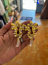 Load image into Gallery viewer, Close-up of a hand holding a pair of ornate gold keyhole-shaped statement earrings with yellow enamel, crystal accents, pearl embellishments, and clear quartz-style drops, photographed inside a boutique with warm lighting and wooden flooring.
