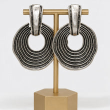 Load image into Gallery viewer, Oversized silver statement earrings featuring layered circular ring detailing and an oxidized textured finish displayed on a jewelry stand.
