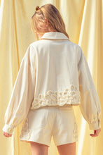Load image into Gallery viewer, Model wearing an écru cropped shirt jacket with oversized floral appliqué embroidery and matching neutral tailored shorts in a soft resort-inspired setting.