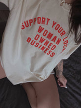 Load image into Gallery viewer, Cream 100% cotton graphic T-shirt with bold red lettering reading "Support Your Local Woman-Owned Business," shown on a model wearing the relaxed-fit tee.