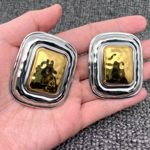 Load image into Gallery viewer, Pair of oversized two-tone statement earrings featuring sculptural rounded rectangular silver-tone frames and raised polished gold-tone centers, displayed in the palm of a hand.