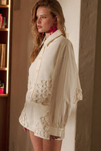 Load image into Gallery viewer, Model wearing an écru cropped shirt jacket with oversized floral appliqué embroidery and matching neutral tailored shorts in a soft resort-inspired setting.