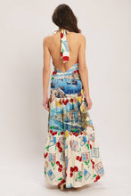 Load image into Gallery viewer, Model wearing a cream halter maxi dress featuring colorful Mediterranean coastal scenes, cherries, lemons, and postcard-inspired prints with a flowing tiered silhouette.
