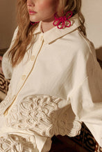 Load image into Gallery viewer, Model wearing an écru cropped shirt jacket with oversized floral appliqué embroidery and matching neutral tailored shorts in a soft resort-inspired setting.