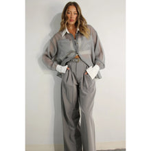 Load image into Gallery viewer, A model showcases a pair of high-waisted, grey pleated trousers, complete with a matching belt, exuding a timeless and sophisticated aesthetic. The trousers feature a tailored fit with front pleats and a straight-leg silhouette, offering both comfort and elegance. Paired with a sheer organza blouse, the ensemble presents a harmonious blend of textures and styles, perfect for both professional and formal occasions.