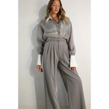 Load image into Gallery viewer, A model showcases a pair of high-waisted, grey pleated trousers, complete with a matching belt, exuding a timeless and sophisticated aesthetic. The trousers feature a tailored fit with front pleats and a straight-leg silhouette, offering both comfort and elegance. Paired with a sheer organza blouse, the ensemble presents a harmonious blend of textures and styles, perfect for both professional and formal occasions.