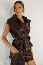 Load image into Gallery viewer, Female model wearing a chocolate brown vegan leather two-piece shorts set featuring a sleeveless trench-style jacket with oversized lapels, a belted waist, and matching shorts.

