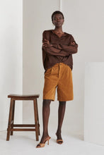 Load image into Gallery viewer, Model wearing high-waisted brown corduroy Bermuda shorts paired with a loose, deep brown blouse and nude heeled mules. She stands confidently next to a wooden stool against a minimal studio backdrop, highlighting the timeless, year-round style of the shorts.

