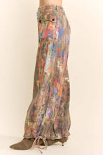 Load image into Gallery viewer, Model wearing abstract print wide leg cargo pants featuring earthy multicolor tones, oversized flap pockets, adjustable tie hems, and a relaxed fit.
