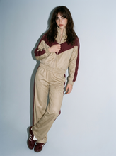 Load image into Gallery viewer, Front view of a female model wearing the Retro Varsity beige and burgundy two-piece track set, featuring a chevron colorblock zip-up jacket and matching high-rise straight-leg pants.
