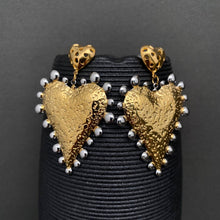 Load image into Gallery viewer, Gold heart-shaped earrings with silver accents on a black stand against a dark background
