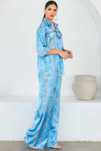 Load image into Gallery viewer, Woman wearing a light blue denim looking satin outfit standing against a white wall with a decorative vase.
