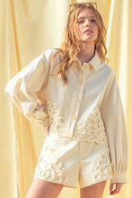 Load image into Gallery viewer, Model wearing an écru cropped shirt jacket with oversized floral appliqué embroidery and matching neutral tailored shorts in a soft resort-inspired setting.
