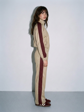 Load image into Gallery viewer, Female model wearing a beige and burgundy retro colorblock track set, featuring a zip-up jacket with burgundy sleeve stripes and matching straight-leg pants with side stripes, shown from the side.
