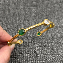 Load image into Gallery viewer, 18K gold-plated open cuff bangle with a hammered textured finish and oval emerald-green and clear zircon stones in gold settings.
