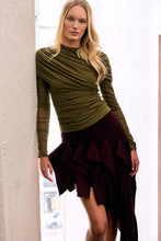 Load image into Gallery viewer, Blonde model wearing an olive green long sleeve mesh top with elegant draped ruching across the bodice and sheer sleeves, styled with a burgundy asymmetric skirt.

