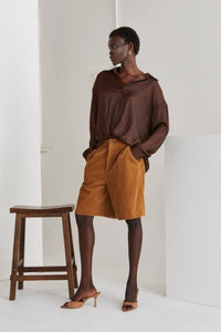 Model wearing high-waisted brown corduroy Bermuda shorts paired with a loose, deep brown blouse and nude heeled mules. She stands confidently next to a wooden stool against a minimal studio backdrop, highlighting the timeless, year-round style of the shorts.
