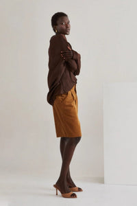 Model wearing high-waisted brown corduroy Bermuda shorts paired with a loose, deep brown blouse and nude heeled mules. She stands confidently next to a wooden stool against a minimal studio backdrop, highlighting the timeless, year-round style of the shorts.