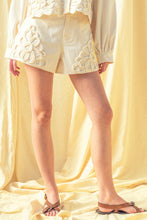 Load image into Gallery viewer, Model wearing écru tailored shorts with oversized floral appliqué embroidery paired with coordinating top in a soft neutral ivory/yellow tone.
