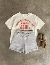 Load image into Gallery viewer, Cream 100% cotton graphic T-shirt with bold red lettering reading "Support Your Local Woman-Owned Business," shown on a model wearing the relaxed-fit tee.
