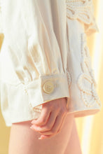 Load image into Gallery viewer, Model wearing an écru cropped shirt jacket with oversized floral appliqué embroidery and matching neutral tailored shorts in a soft resort-inspired setting.
