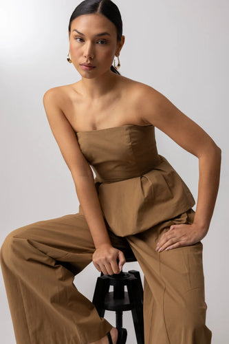 Woman wearing a camel toned strapless top and wide-leg pants sitting on a black stool against a light gray background.