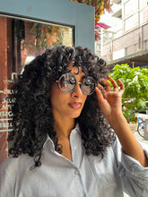 Load image into Gallery viewer, Model wearing oversized round black sunglasses with UVA and UVB protection, standing in front of Glam Expressway Boutique in DUMBO Brooklyn. The sunglasses feature dark gradient lenses and bold frames, styled with a light pinstriped button-down shirt and voluminous curly hair. A fashionable summer accessory perfect for city chic and sun-safe style.
