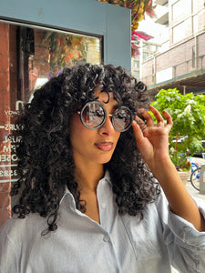 Model wearing oversized round black sunglasses with UVA and UVB protection, standing in front of Glam Expressway Boutique in DUMBO Brooklyn. The sunglasses feature dark gradient lenses and bold frames, styled with a light pinstriped button-down shirt and voluminous curly hair. A fashionable summer accessory perfect for city chic and sun-safe style.
