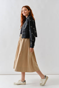 Woman wearing a Glam Expressway black leather jacket with grommets, beige skirt, and white sneakers on a white background