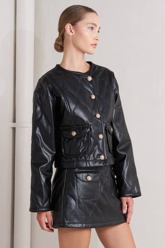 Woman wearing a Glam Expressway black leather jacket and skirt set against a plain background