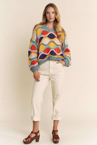 Woman wearing a colorful patterned sweater and white pants against a plain background