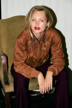 Load image into Gallery viewer, Woman wearing a brown suede jacket with gold accents sitting on a chair.
