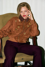 Load image into Gallery viewer, Woman in a brown jacket and purple pants sitting on a patterned chair.
