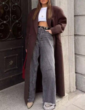 Load image into Gallery viewer, Person wearing a long brown coat, white top, and gray jeans standing against a building.
