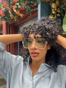 Model wearing oversized square green sunglasses with transparent frames and tinted lenses, posing in front of Glam Expressway Boutique in DUMBO, Brooklyn. Trendy summer eyewear offering UV protection and bold retro style.