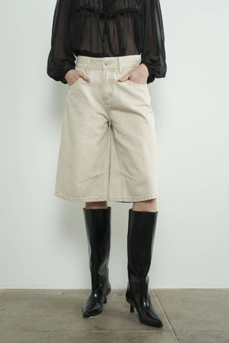Person wearing a black sheer top, beige mid-waisted cream denim bermuda shorts, and black knee-high boots against a white background.