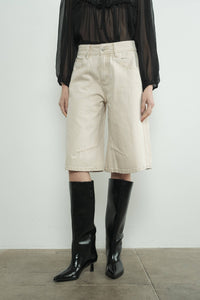 Person wearing beige denim bermuda shorts and black knee-high boots on a plain background