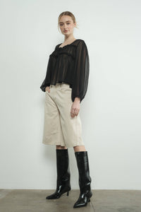Woman wearing a black blouse, beige denim bermuda shorts, and black knee-high boots against a white background