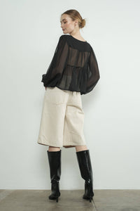 Woman wearing a black sheer top, beige bermuda shorts, and black boots against a white background