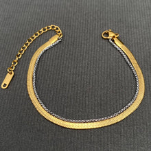 Load image into Gallery viewer, Two-tone layered bracelet featuring a flat 18K gold-plated herringbone chain alongside a delicate silver-tone link chain, with lobster clasp and adjustable extender, displayed on gray fabric.
 
